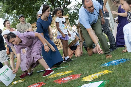A crowd of people , standing and crouching, play a game with giant playing cards in the grass.