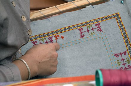 Closeup of someone embroidering a colorful border on pale blue fabric sewn to a wooden frame.