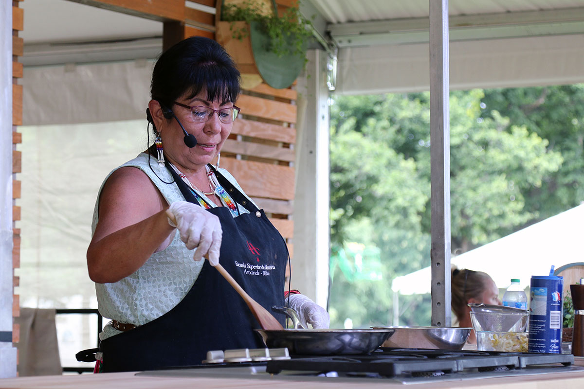 A woman wearing a headset microphone and a black apron stirs something in a pan on a stove in an outdoor demonstration kitchen.
