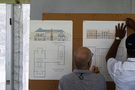 Backshot of two men work together to pin blueprints of a building design to a corkboard wall.