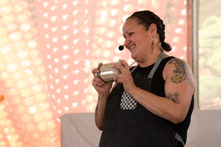 A woman wearing a black tank top and apron holds up a jar of grain, with a photo backdrop of ears of red corn.