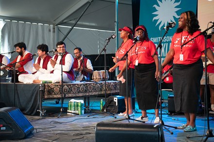 Two musical groups on stage together, one group in white with red vests, seated on a riser, and the other standing in matching black pants and red shirts.