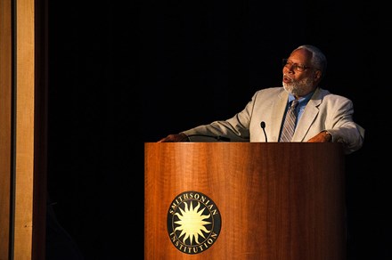 A man in a gray suit speaks on a stage at a podium with the Smithsonian sun logo in gold and black.