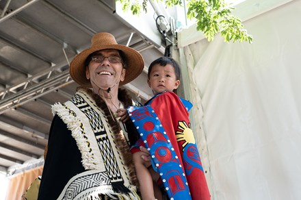 A man wearing a black and white woven shawl and a woven brimmed straw hat smiles, holding up a toddler in a similar shawl but with colors and designs of the Philippines flag.