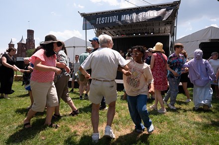 On a patch of grass in front of a stage, pairs of two people have their arms linked and spin around in circles. In the foreground, an older man and younger girl dance together.