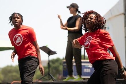 Two young performers dancing and wearing red t-shirts designed with a white logo for Stax Music Academy’s 25th anniversary; a person stands onstage behind them, translating to American Sign Language for an off-camera audience.