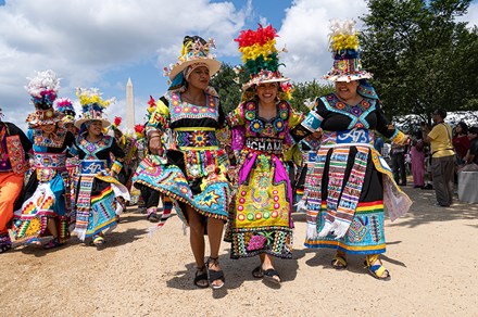 A group of people in bright and colorful traditional Bolivian clothing and hats process down the National Mall with their arms around each other, kicking their feet up, big smiles on their faces.