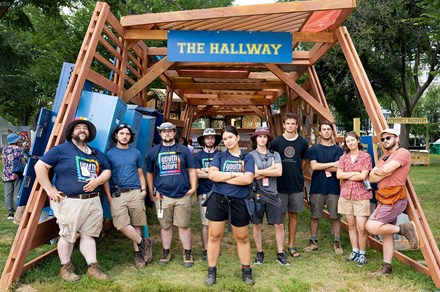 Ten members of the 2025 Smithsonian Folklife Festival's tech crew posing under the framing of The Hallway's wooden structure; six have their arms crossed, two in their pockets, and two by their sides.