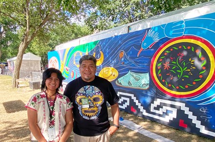 A man and a woman smile in front of an outdoor mural, depicting a leather sandal, flowers, a bird, gold jewelry, a brimmed hat, and other symbols on a dark blue background.