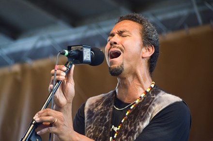 A man with short black hair and goatee, wearing an animal-skin vest and beaded necklace, sings into a microphone on stage.