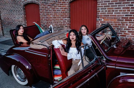 Three women with long dark hair pose in a shiny, port wine-colored classic convertible car with the top down.