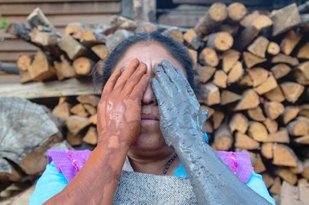 A woman holds her hands over her face, covering her eyes. Her hands and bare forearms are covered in clay: one side red, one side bluish gray. Behind her are stacks of chopped wood.