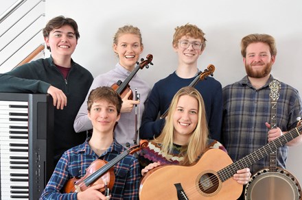 Six young people pose, smiling, in two rows with their instruments: electric keyboard, fiddles, banjo, and acoustic guitar.
