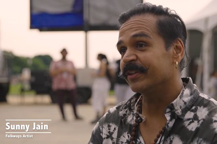A South Asian man with black handlebar moustache, gold earring, and tie-dyed shirts speaks, seated. HIs name is in text: Sunny Jain, Folkways Artist. Behind him, backstage of an outdoor festival.
