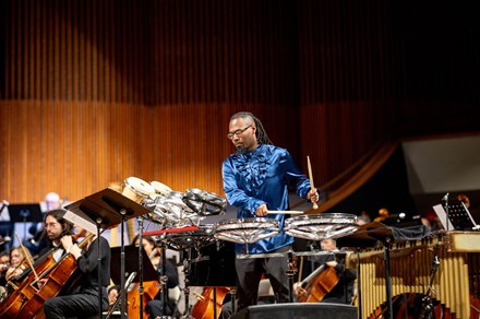 A man in an iridescent blue dress shirt plays drums on stage with an orchestra behind him.