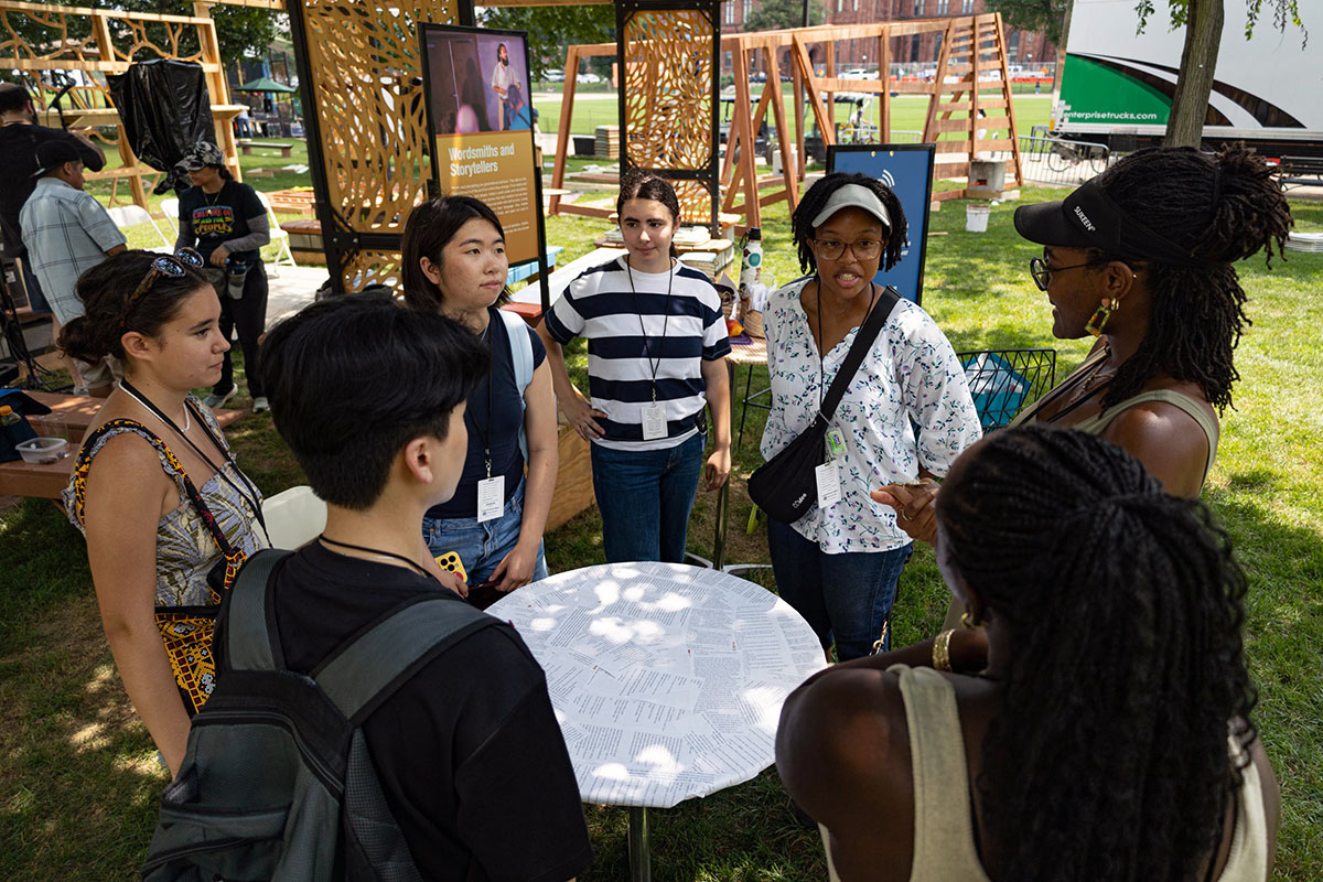 Seven people gather in a circle around a coffee table collaged with pages of text. Wooden festival structures are going up behind them.
