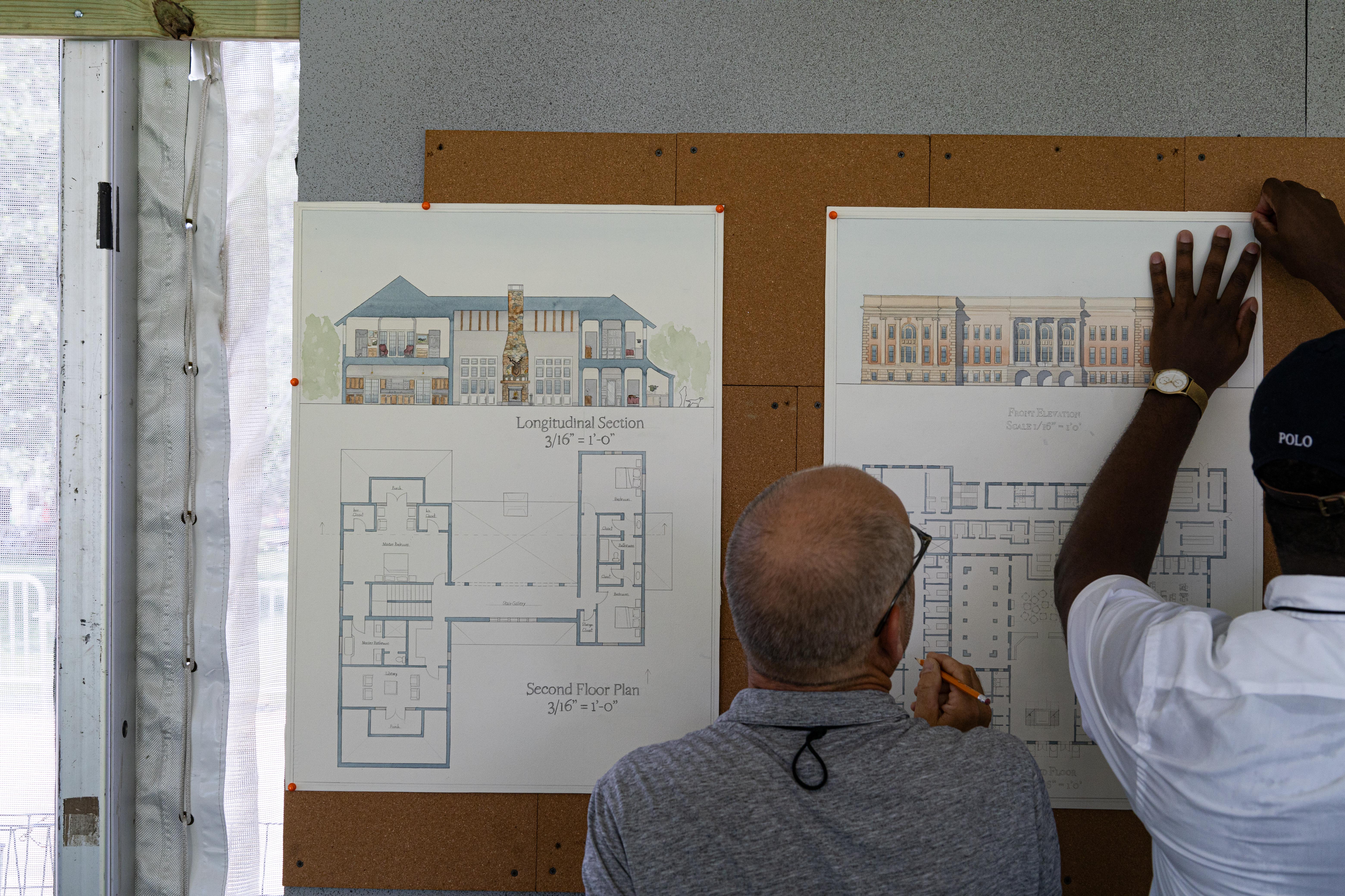 Backshot of two men work together to pin blueprints of a building design to a corkboard wall.