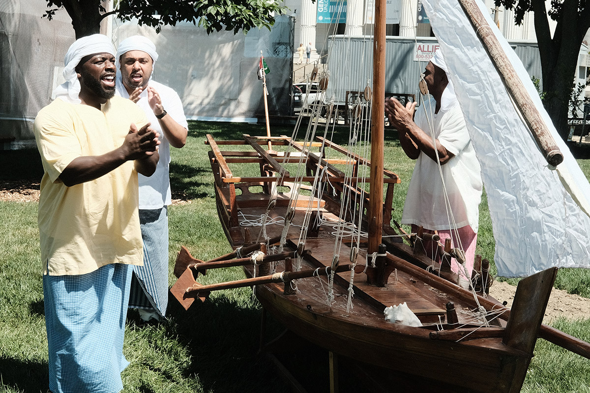 Three men dance and clap around a small wooden boat with a white sail.