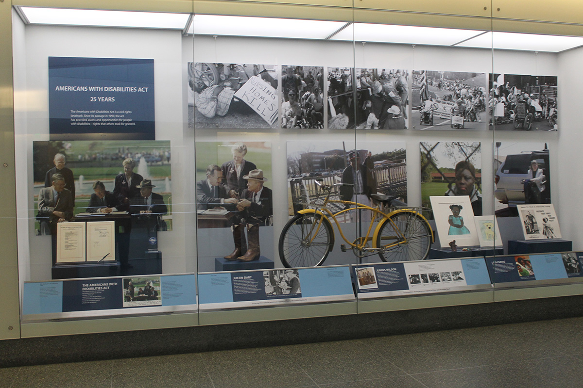A large museum display case, lit from above and behind glass. A sign at the top reads AMERICANS WITH DISABILITIES ACT / 25 YEARS, but the blurb below is too small to read. Items in the case include blown-up photos of protests in black and white, President George H.W. Bush signing the act into law, an old yellow bicycle, and a timeline.