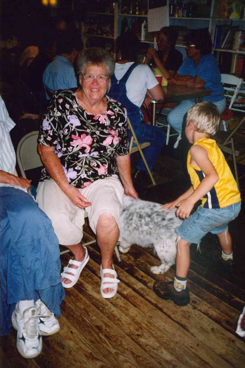 A seated woman with short gray hair smiles at the camera as she and a child, faced away, pet a dogs. In the background, musicians sit in a circle.