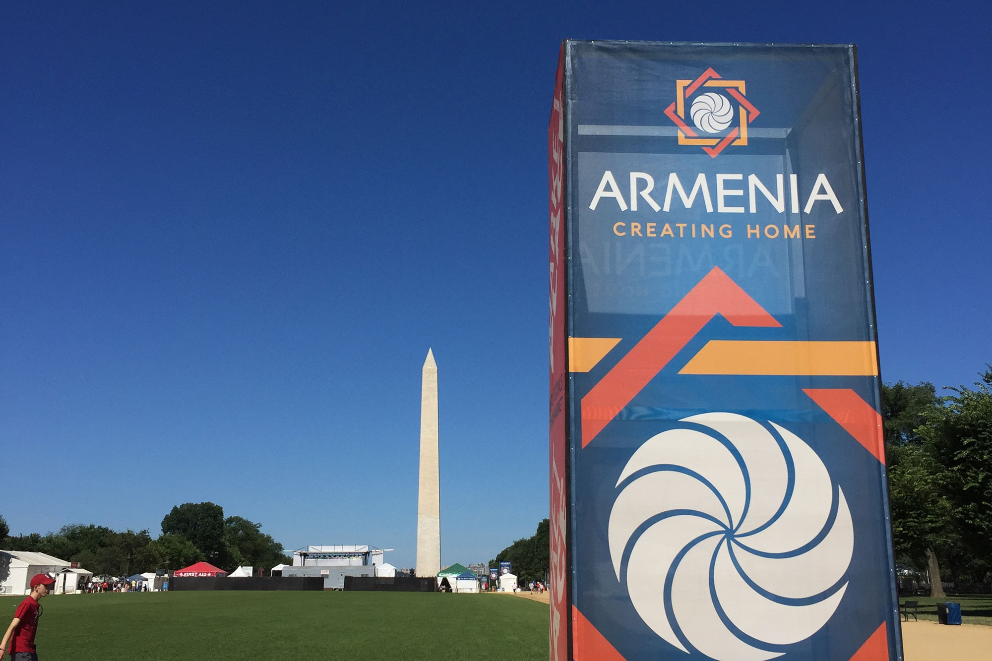 In the left foreground, a colorful sign with the words “Armenia Creating Home” stands on a patch of grass next to a dirt path. A large, white obelisk points towards the blue sky in the background. Between the sign and the obelisk is a large green field, with some tents and a sound stage.