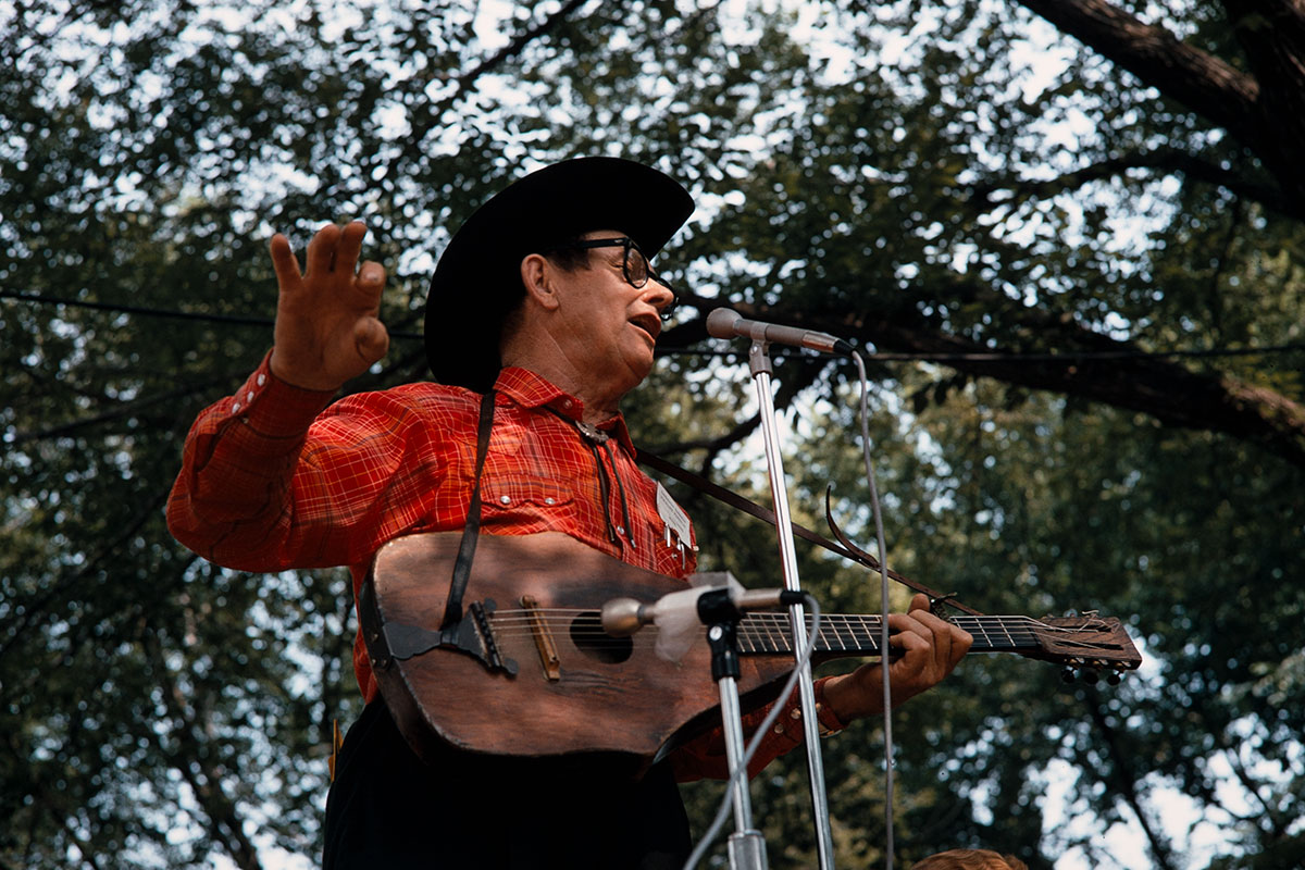 A man in a black hat, black glasses, and a red plaid shirt performs under trees, strumming an oddly shaped wooden guitar.