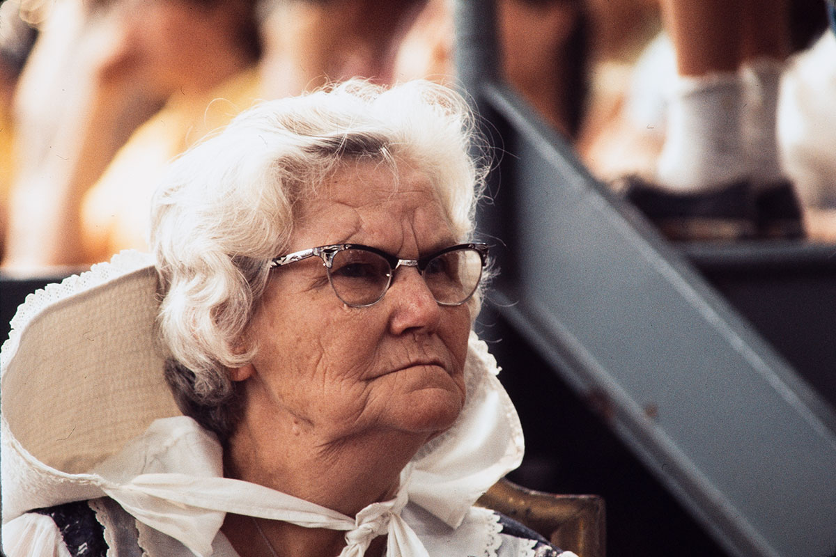 An elder white woman with short, wavy white hair, horn-rimmed glasses, and a white bonnet tied at her neck looks on sternly.