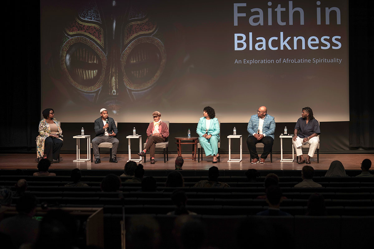 Six people seated on stage before a seated audience. On the projection screen behind them, a close-up on the eyes of a painted  mask and the discussion title: Faith in Blackness: An Exploration of AfroLatine Spirituality.