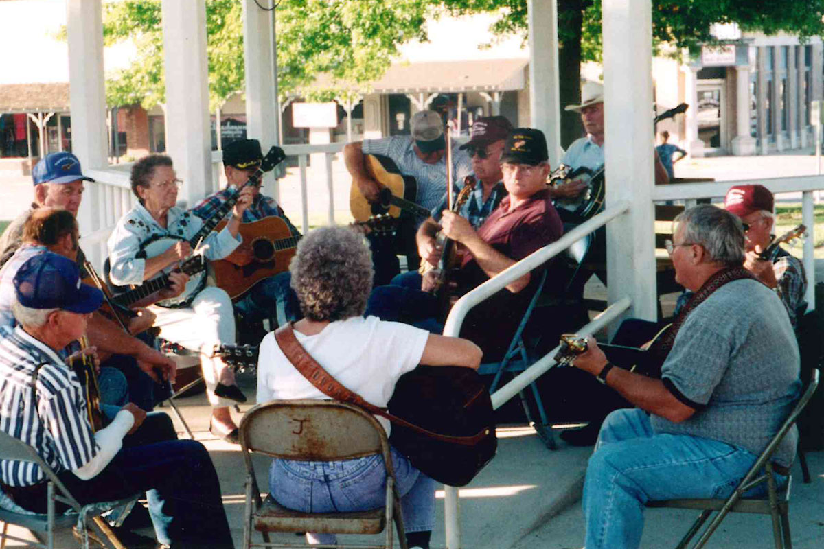 About a dozen elder people sit in a circle of folding chairs under a white gazebo playing acoustic guitars, banjos, and fiddles. 