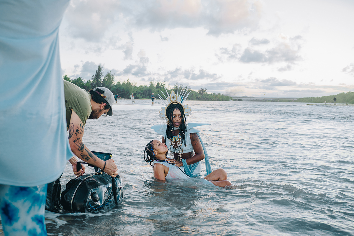 One person in fantastical dress, including a radiating headdress and long, exaggerated shoulder epaulettes, holds another person at the surface of a body of water. A man with tattoos on his arms films them with the camera in a waterproof bag.