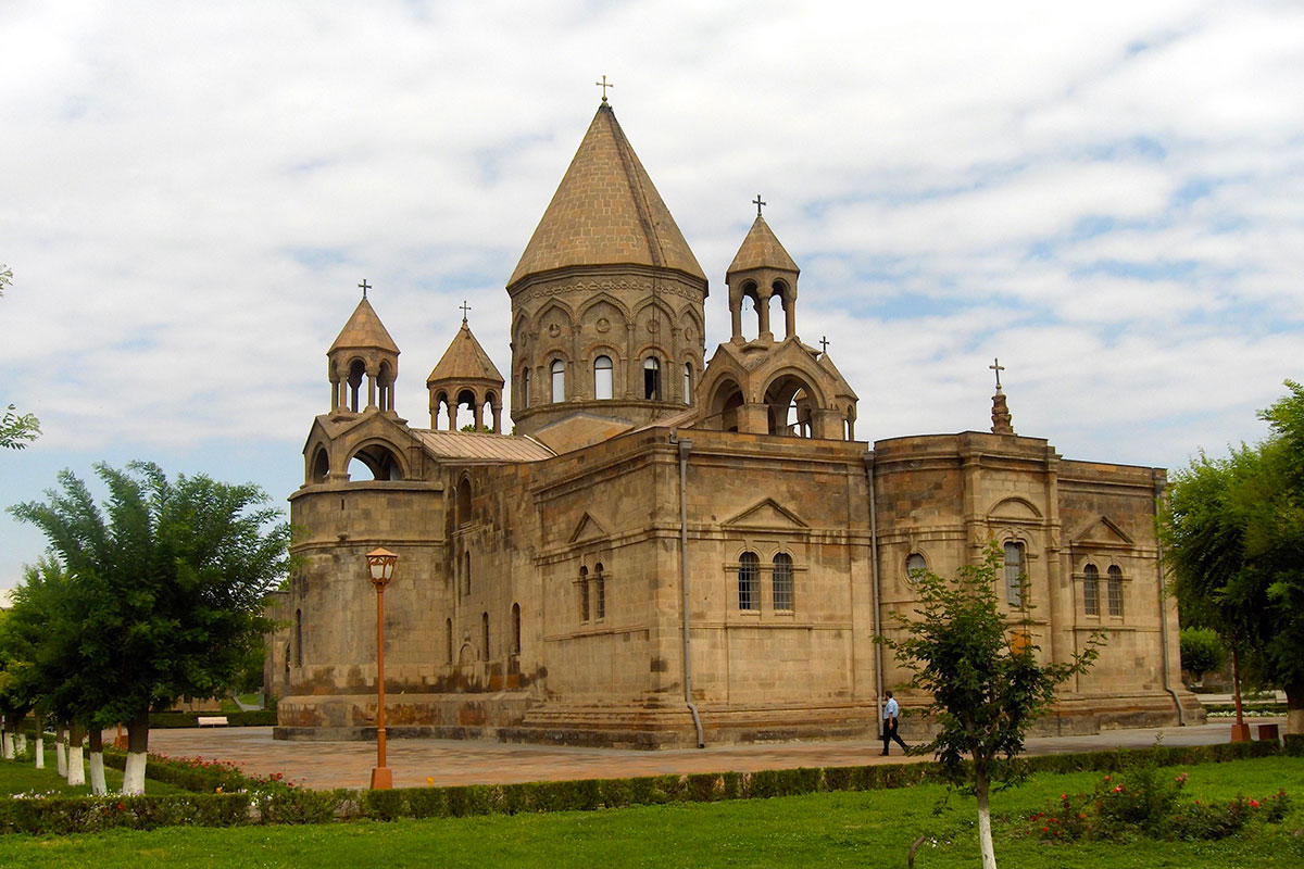 A beige stone church building surrounded by green trees and lawn.