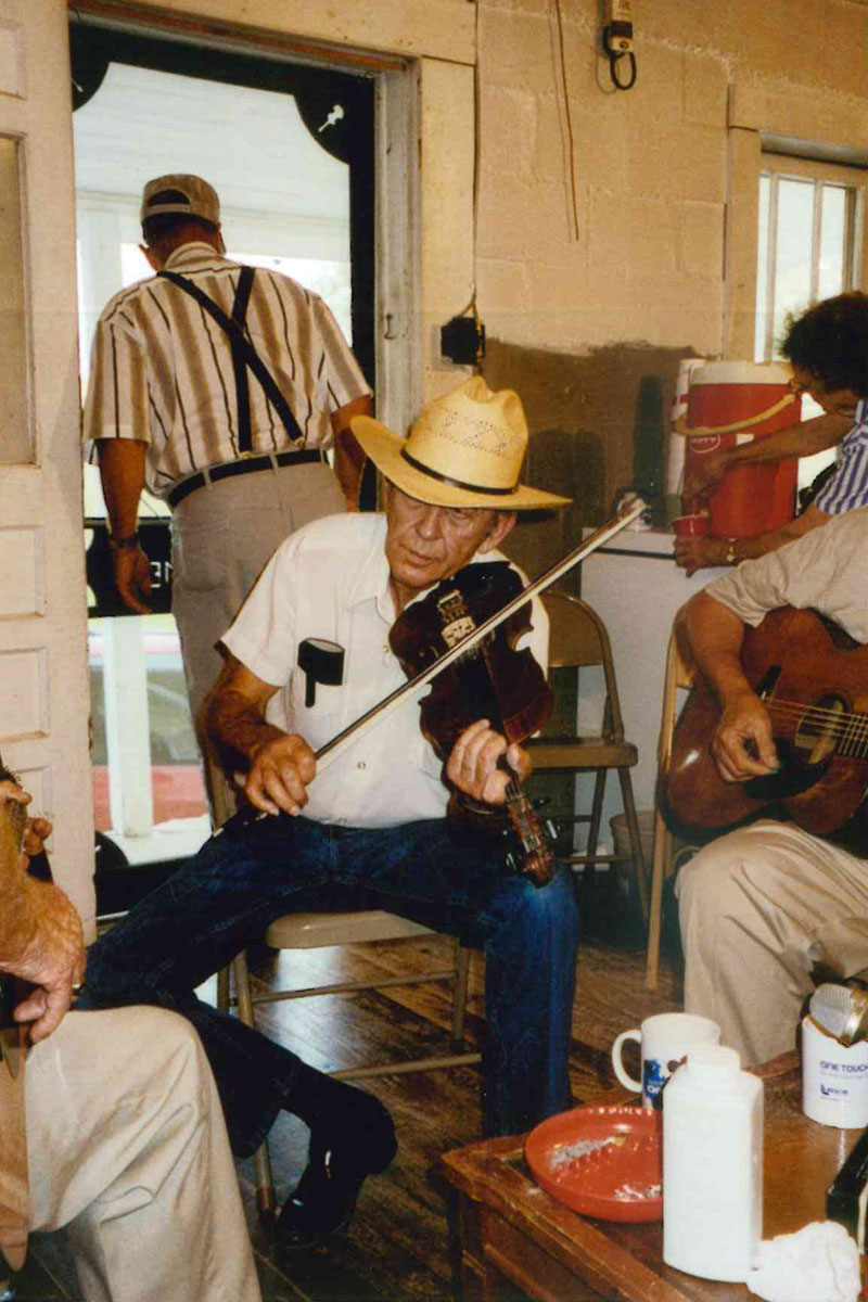 A seated man with a cowboy hat plays fiddle with other musicians cut out of frame in a home.