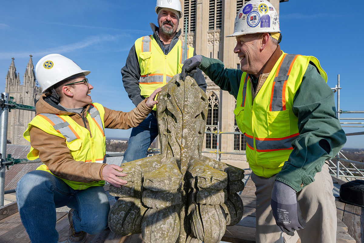 Three people in yellow safety vests and white hard hats smile atop a stone cathedral, with their hands on a decoratively carved piece of stone.
