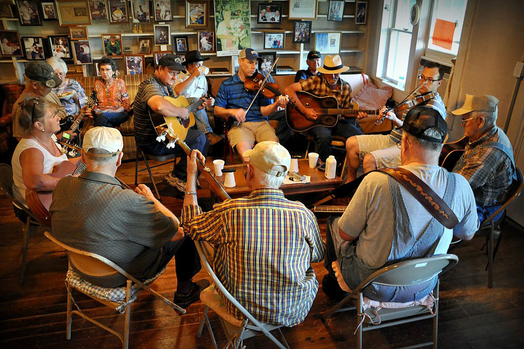 About 14 people sit in a circle indoors playing acoustic guitars and fiddles.