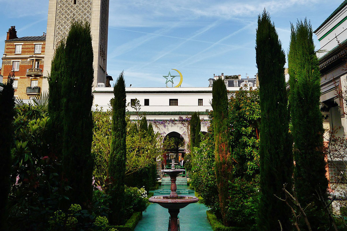 A courtyard with a small  pool and water fountain in center, flanked by trees and other plants. In the background, a building with an arched entrance and Islam star and crescent moon mounted on the roof.