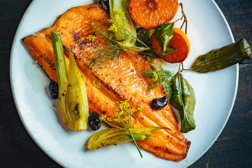 Pink fillet of fish on a round white dish, garnished with green onions and herbs and orange citrus halves.