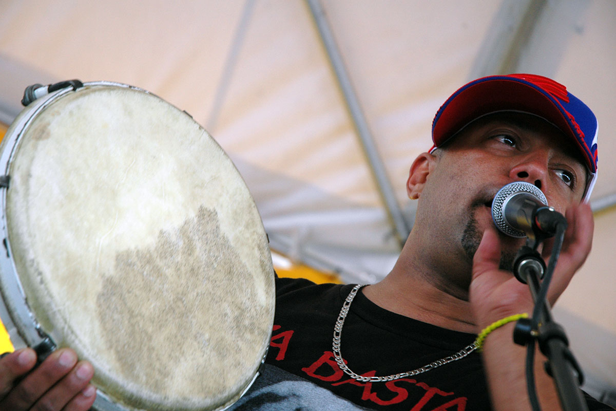 A man with ballcap and chain necklace holds a hand drum in one hand and a microphone in the other as he speaks into it.