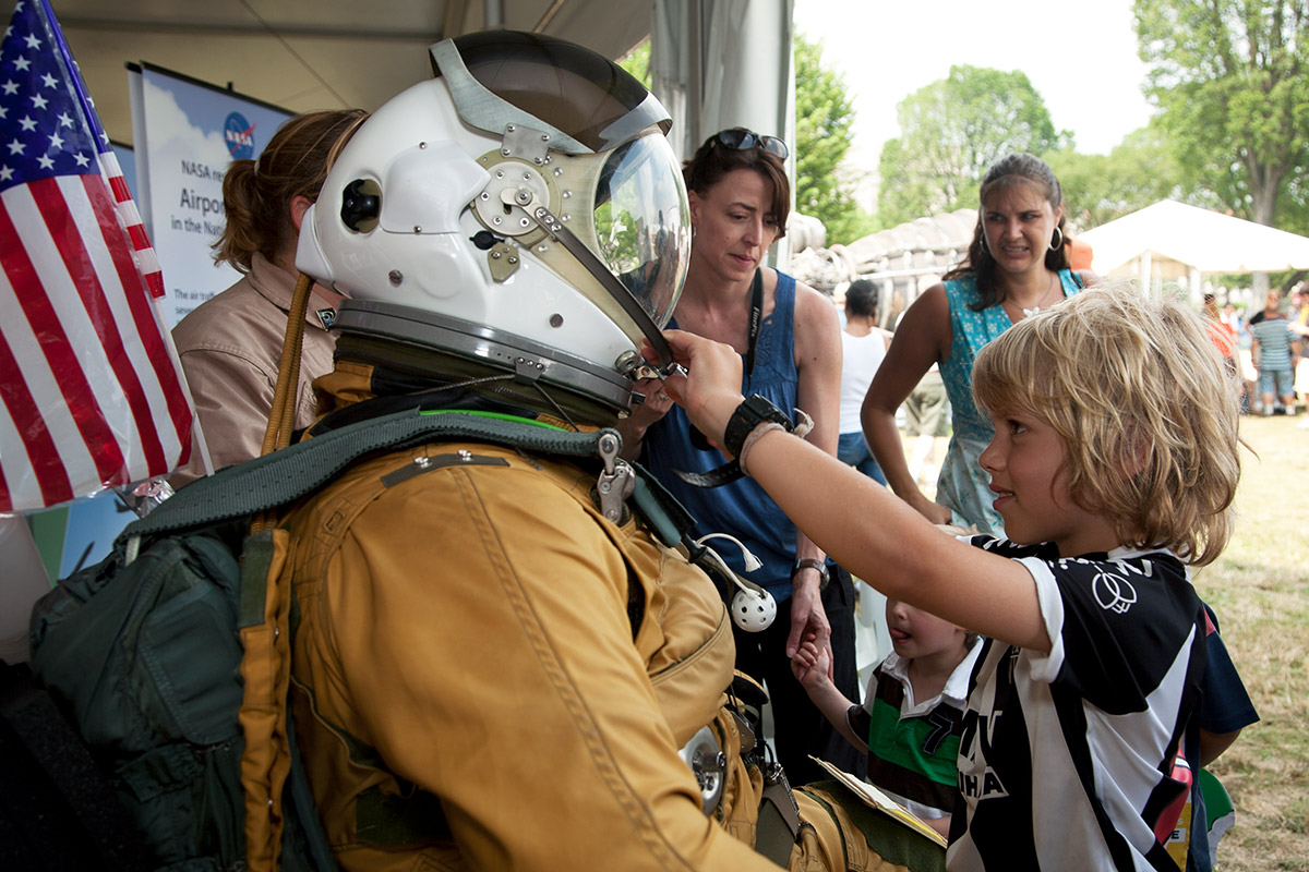 A small child grips the helmet of an astronaut suit, partially covered by a tent. A collection of adults and children look on in the background. 