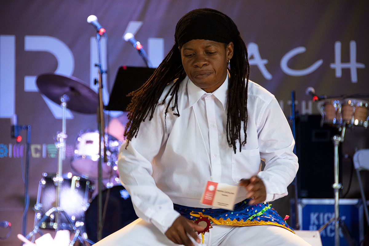 A Black woman dressed in white dress shirt and pans, black headband, and blue and gold sash belt, with long dark locs, performs on stage.