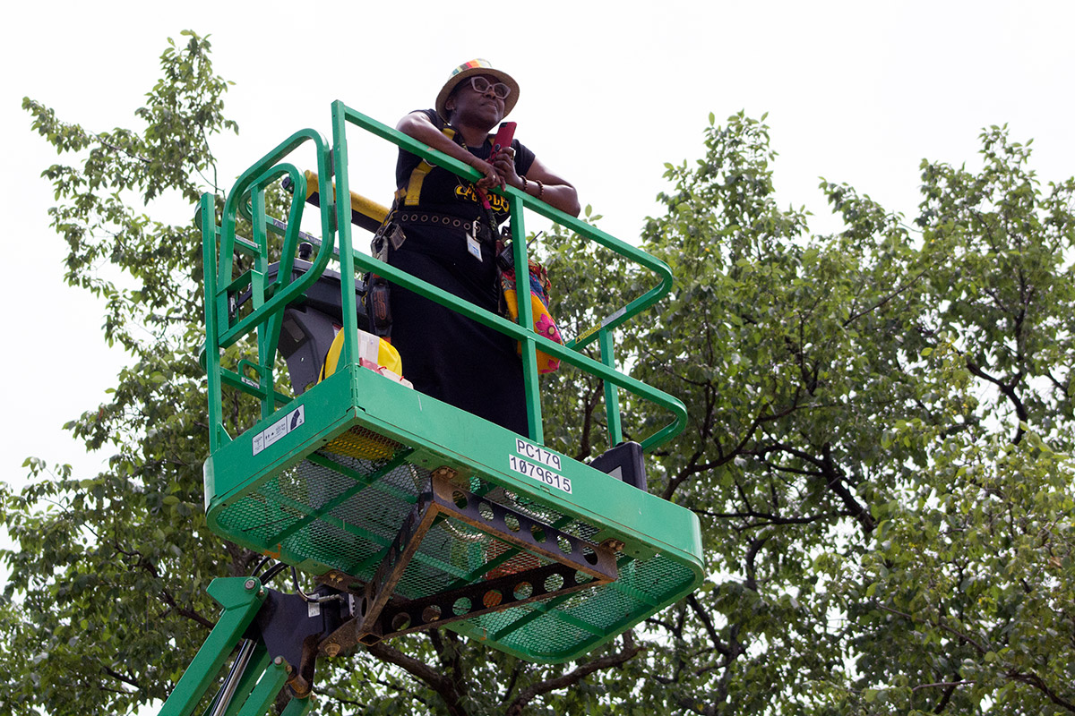 A Black woman standing on a green cherry picker, bright white sky and green trees behind her.