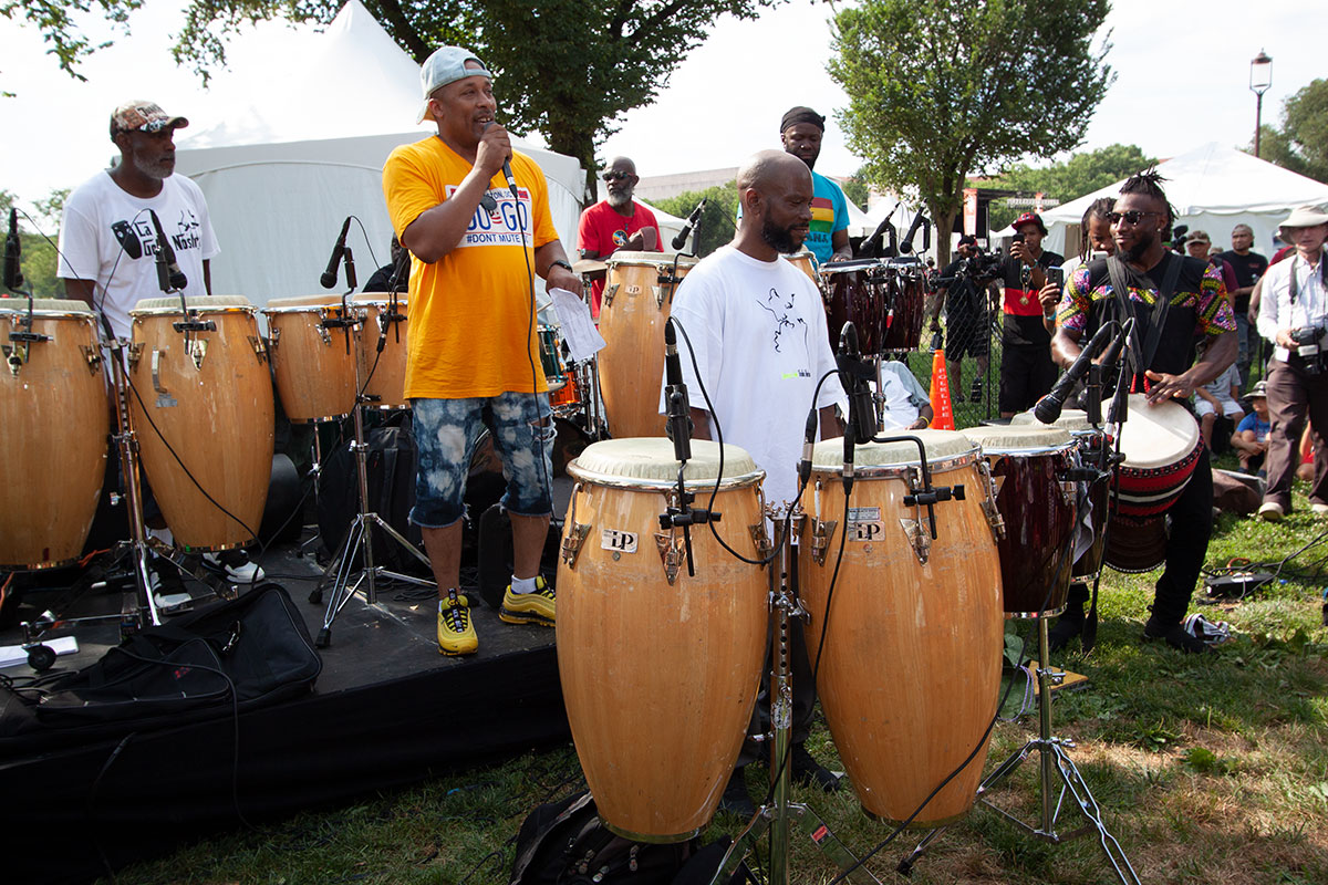 Man in bright yellow shirt addresses an outdoor crowd on microphone as other men on congas around him prepare to perform.