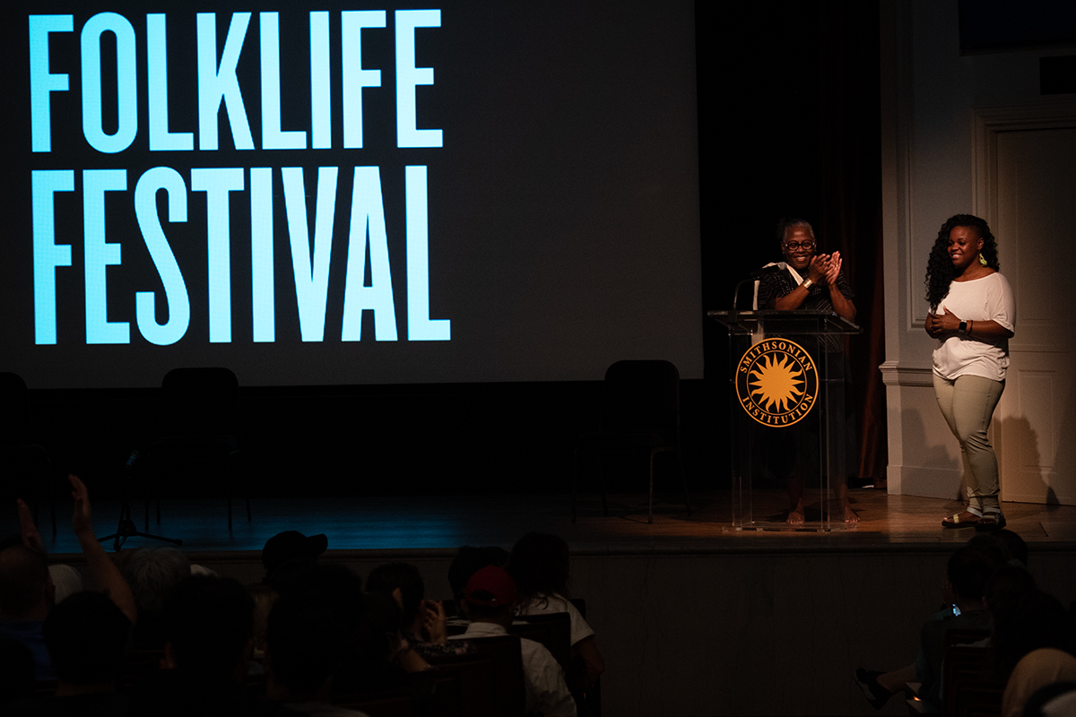 On a dimly lit stage, one woman stands behind a podium with the Smithsonian sunburst logo, with another beside her. On the projection screen to their right, large white text reads FOLKLIFE FESTIVAL.