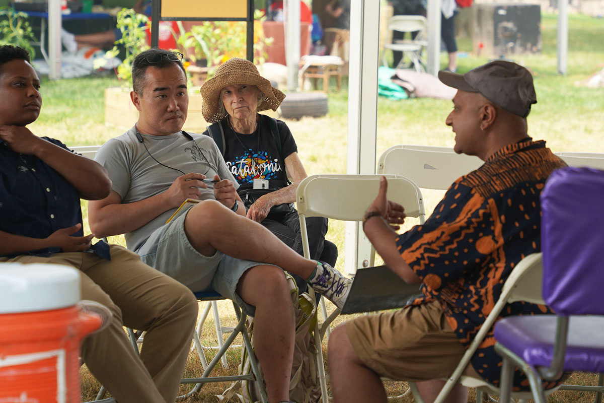 A man sits opposite three people under a tent outdoors, all in folding chairs.