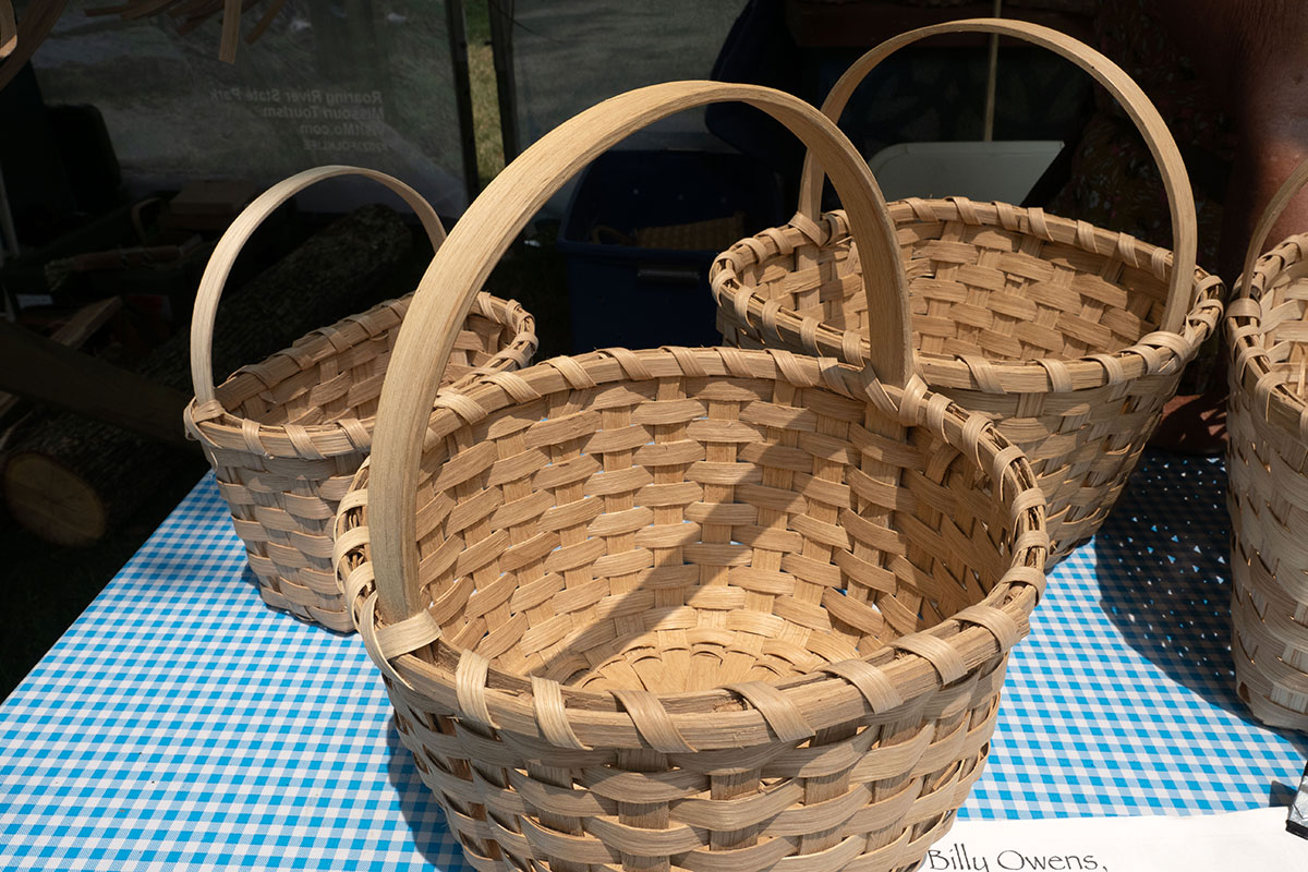 Three tan colored, circular woven baskets, each with an arc-shaped handle, displayed on a gingham tablecloth.