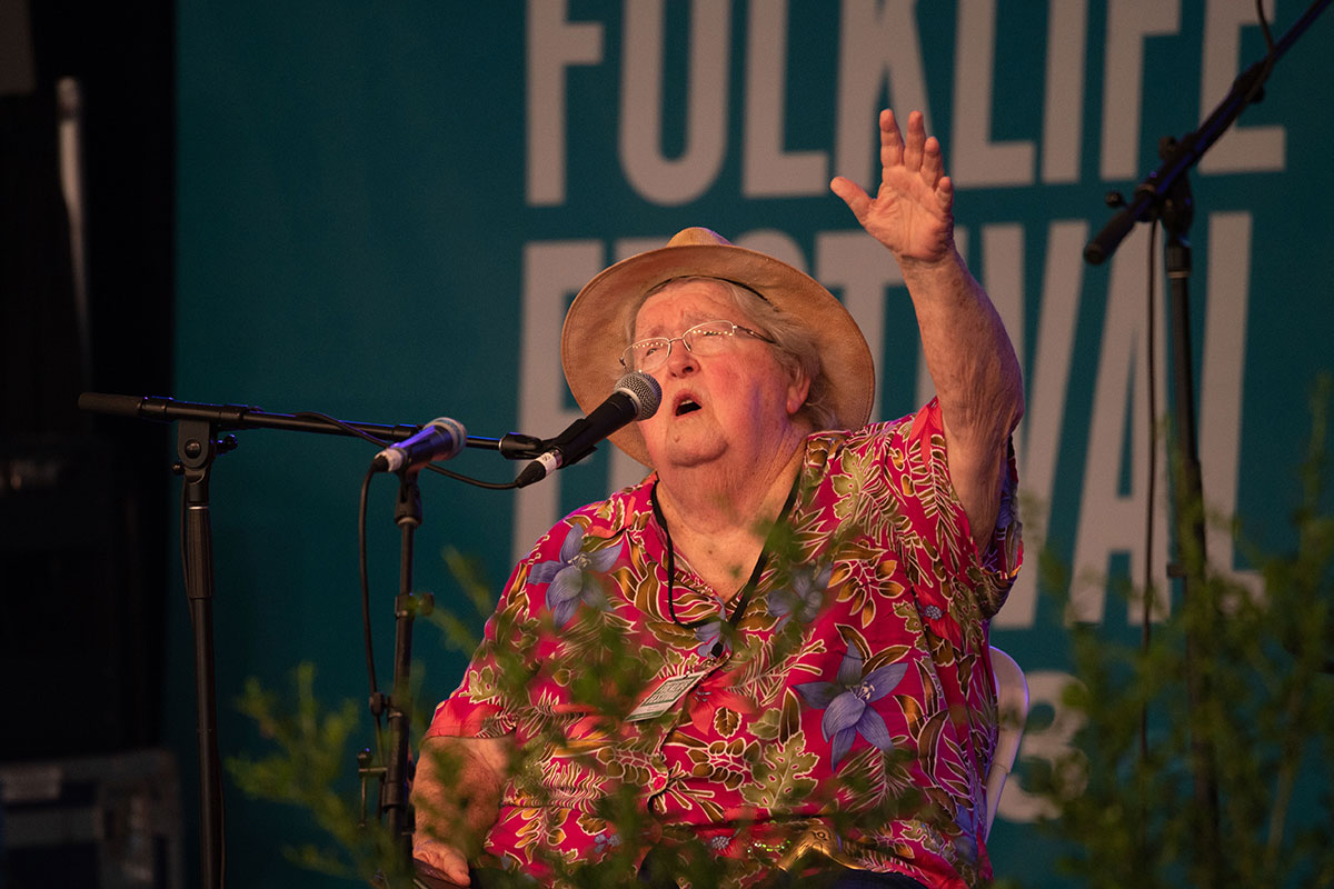 A woman in a red floral blouse and brimmed hat, seated on stage, raises one arm as she sings.
