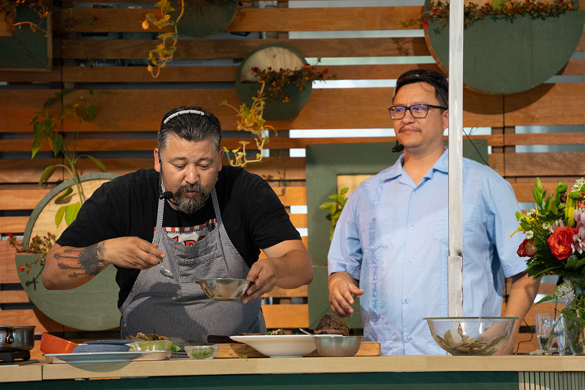 Two men stand at a kitchen counter. One, wearing an apron and a headset microphone, holds a silver bowl and a spoon.