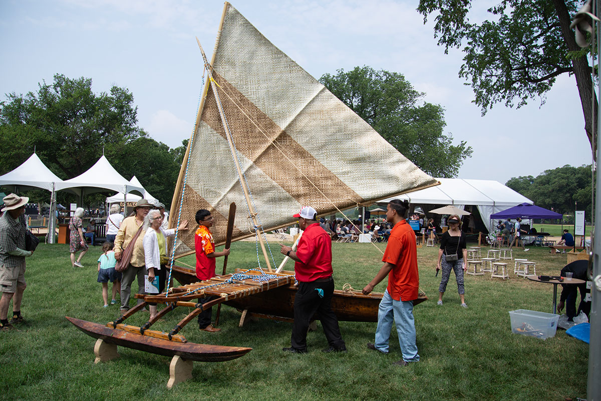 Three men in red  shirts plus visitors stand around a wooden outrigger canoe set up on grass. The woven triangular sail is raised.