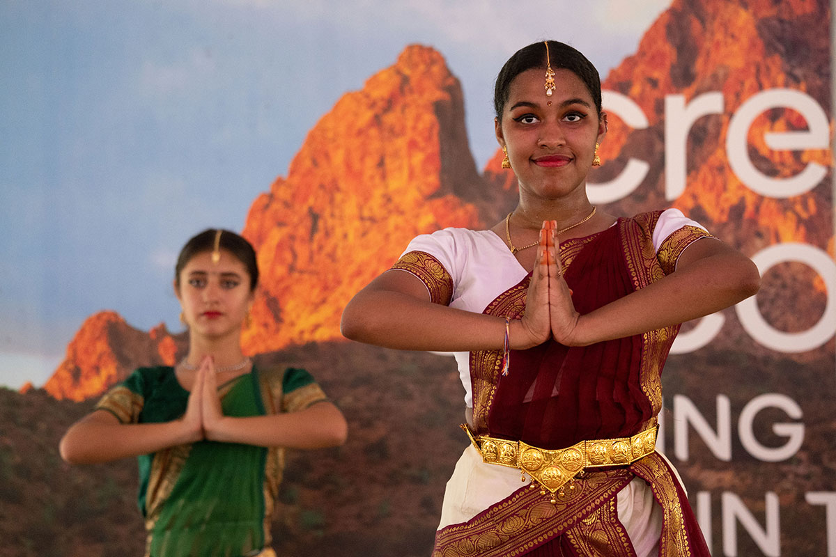 Two dancers on stage, wearing gold-fringed dresses and jewelry, pose with their palms together as if in prayer.