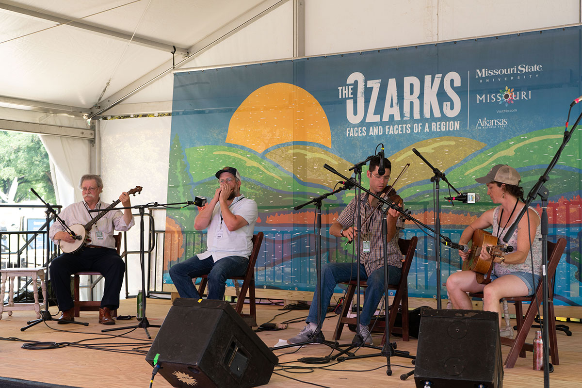 Four musicians perform seated on an outdoor stage: a banjo player, harmonica player, fiddle player, and acoustic guitarist.