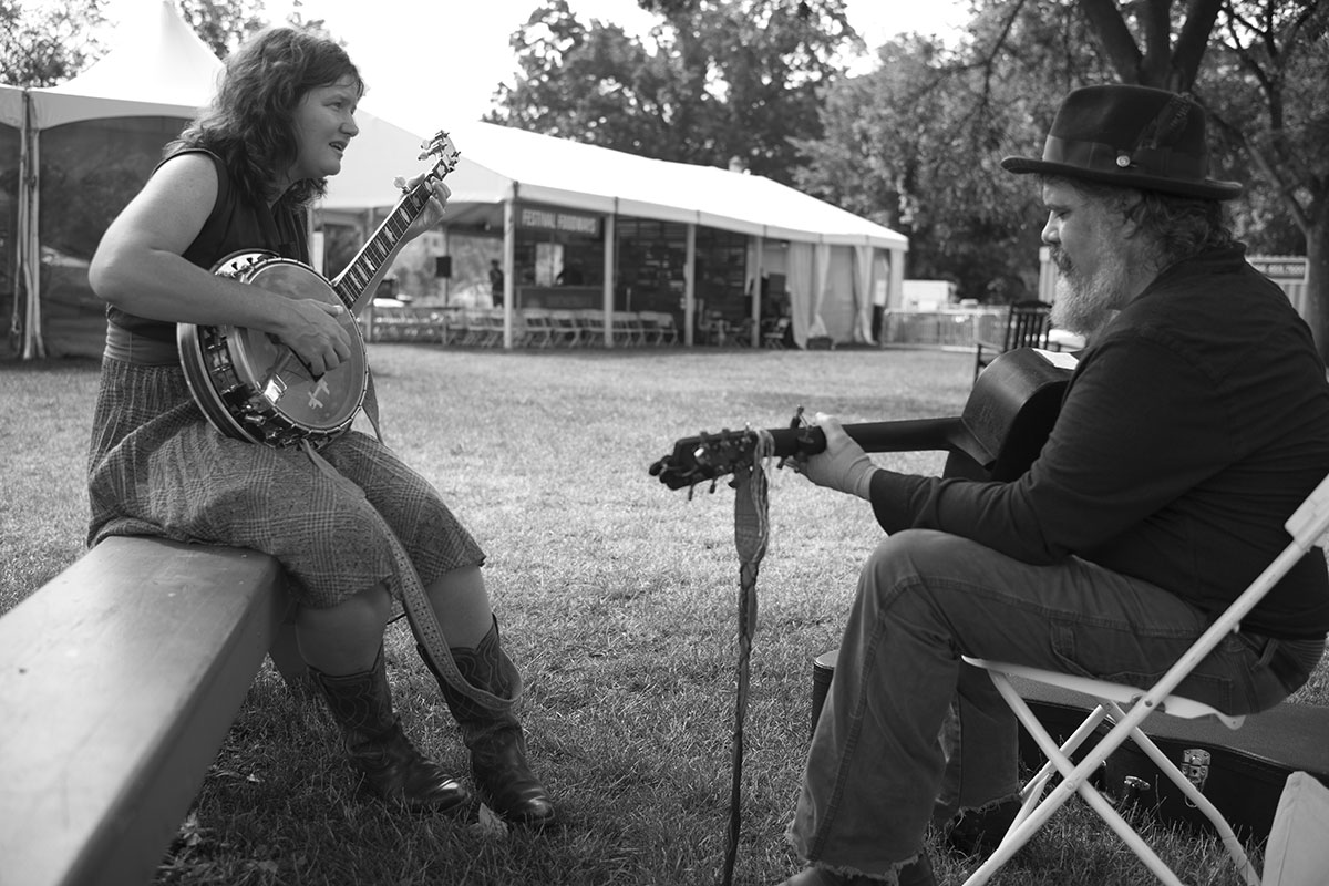 Facing each other, a woman plays banjo and a man plays acoustic guitar outdoors. Black-and-white photo.